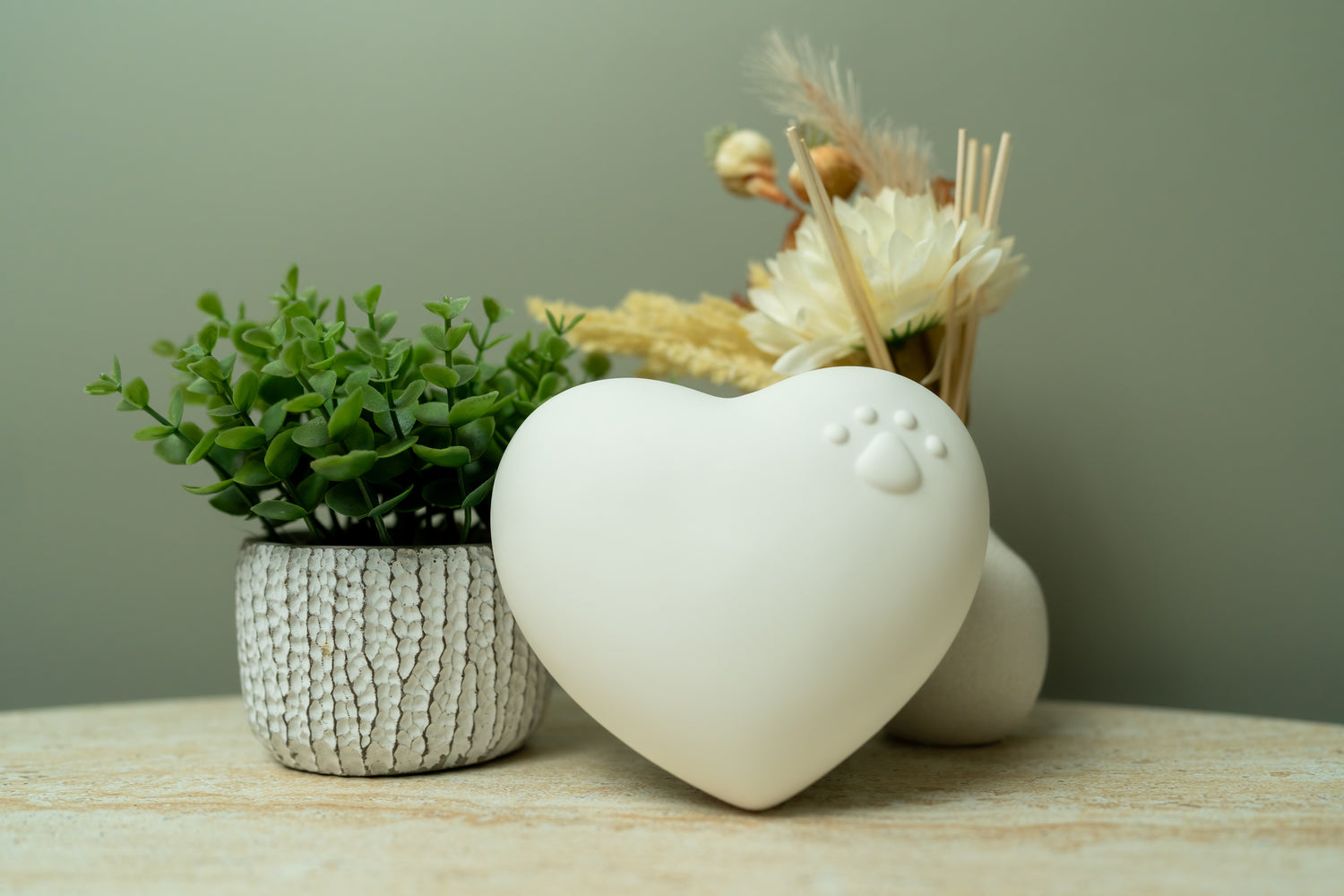A matte white, heart-shaped pet urn with a raised paw print rests on a light wooden surface. To the left is a small potted green plant in a textured white pot, and to the right is a minimal ceramic vase with dried flowers and reed sticks. The soft green wall in the background and natural decor create a warm, peaceful, and elegant memorial setting.