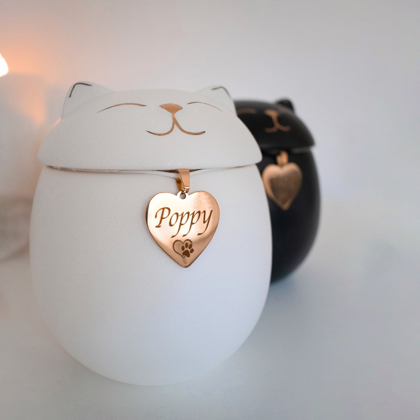 Two cat-shaped pet urns with peaceful facial features, one white and one black, are displayed on a white surface. Each urn has a shiny gold heart-shaped name tag hanging from the neck area. The black urn is engraved with the name "Misty" and the white urn is engraved with the name "Poppy".