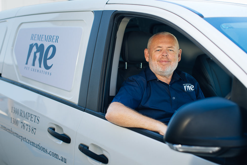 Gary Ditchfield, wearing a navy blue "Remember Me Pet Cremations" shirt, sits in the driver’s seat of a white company van with the business logo displayed on the side. The van also shows the contact number and website for the pet cremation service. Gary smiles warmly, conveying a sense of compassion and professionalism.