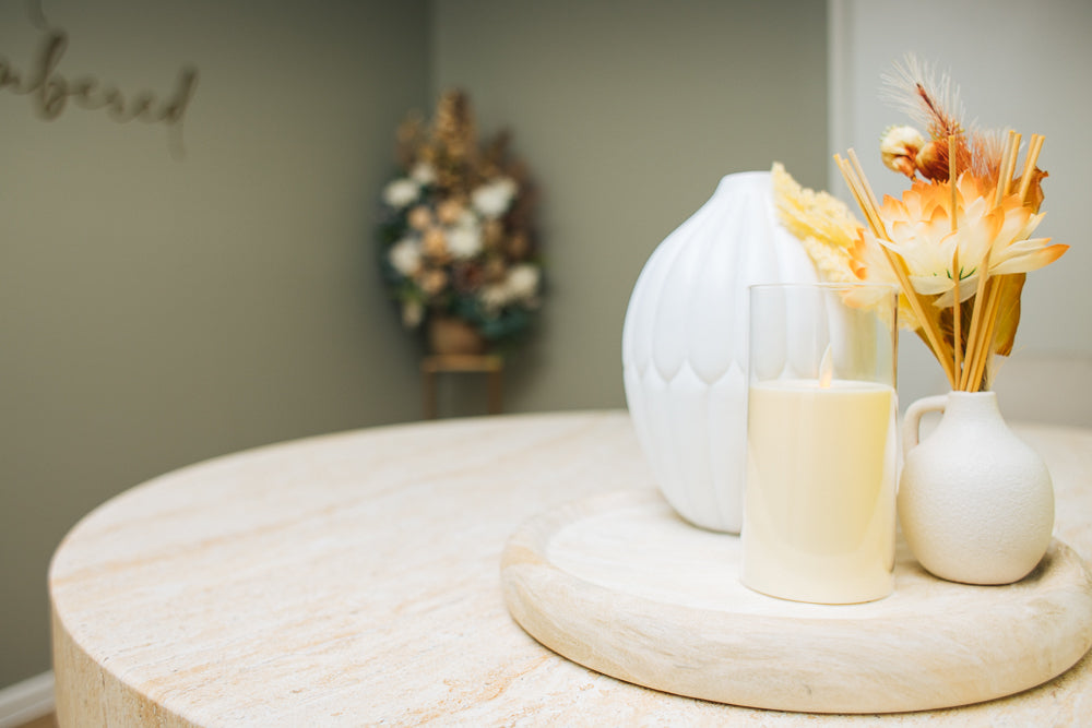 A peaceful setting featuring a round stone table adorned with a soft cream tray holding a white candle, a small vase of dried flowers, and a decorative ceramic piece. In the softly blurred background, a floral arrangement rests against a muted green wall with the word "remembered" partially visible.
