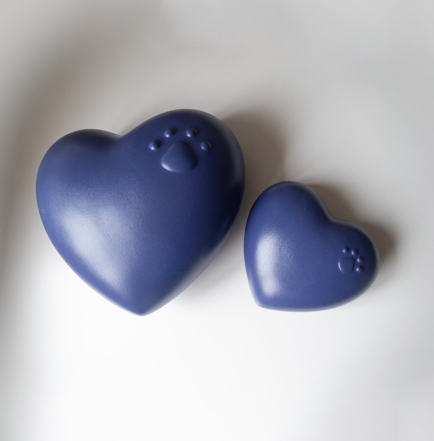 Two heart-shaped pet urns in a matte deep blue finish placed on a white surface. The larger urn features an embossed paw print with four toes, while the smaller urn has a similar paw print design. The shapes and smooth finish give a soft, comforting appearance, suitable for memorialising a beloved pet.