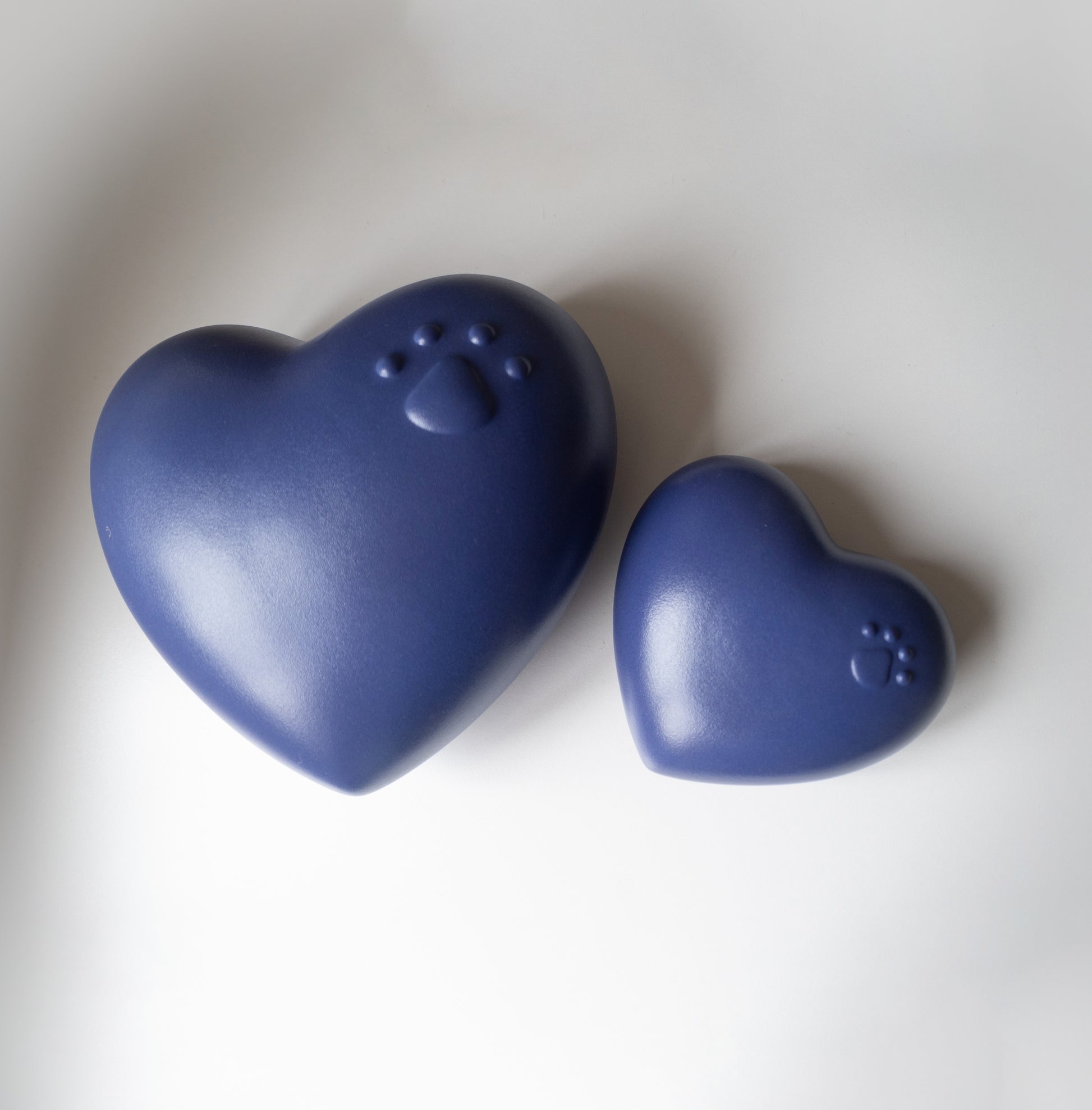 Two heart-shaped pet urns in a matte deep blue finish placed on a white surface. The larger urn features an embossed paw print with four toes, while the smaller urn has a similar paw print design. The shapes and smooth finish give a soft, comforting appearance, suitable for memorialising a beloved pet.