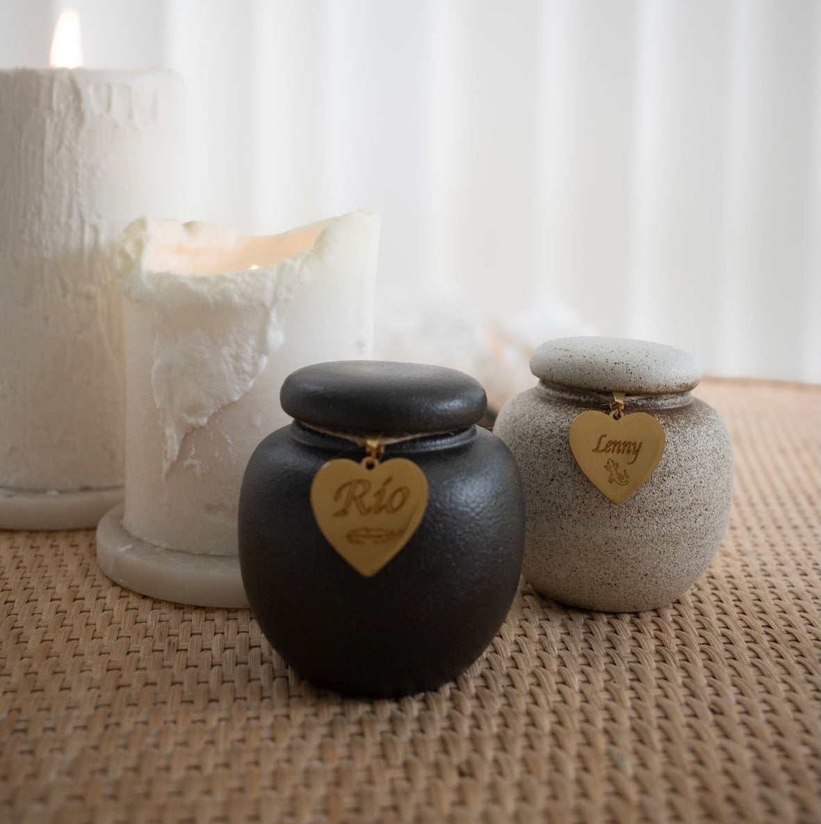 Two small cremation urns sit side by side on a woven surface, each with a gold heart-shaped tag—one engraved with "Rio" and the other with "Lenny." Behind them are softly glowing white candles, contributing to a peaceful and respectful atmosphere.