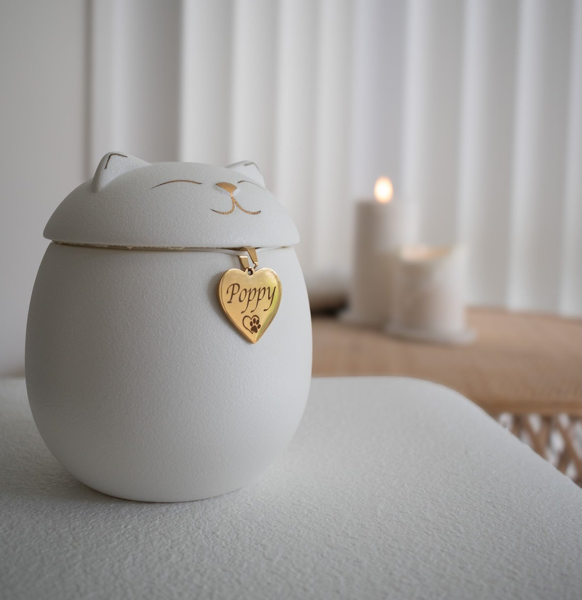 A white ceramic cat-shaped urn with a peaceful face and pointy ears sits on a soft surface. A gold heart-shaped tag engraved with the name "Poppy" and a paw print hangs from the lid. In the softly lit background, two white candles add a warm, calming ambiance.